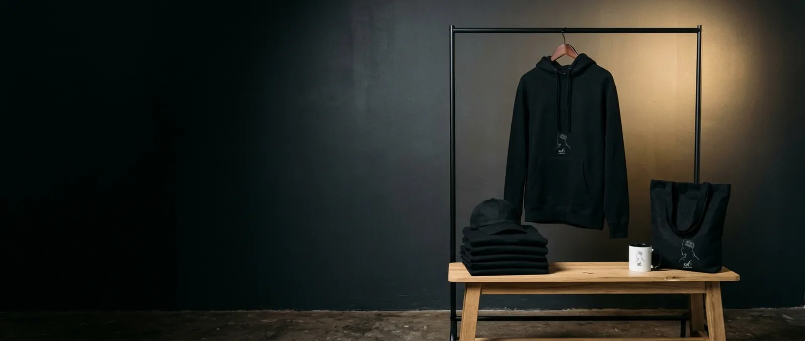 A Kufi Productions hoodie on a rail, with a tote bag, a mug, a cap and folded tees on a wooden bench